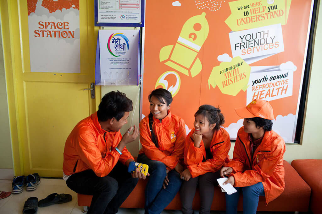 Four young Nepalese people in orange jackets sit and talk in front of a sign that reads "Youth sexual reproductive health".