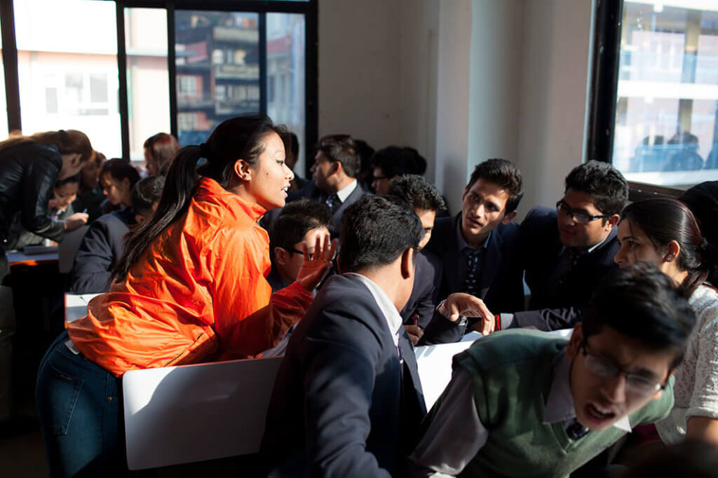 A young woman in an orange jacket talks to a group of young men about youth sexual health.