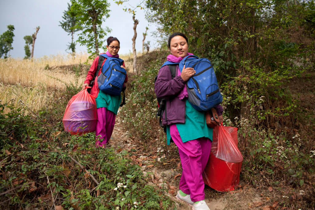 Two nurses carrying medical gear travel down a mountain path. As private providers, they fill the gaps left by Nepal's universal health care system.