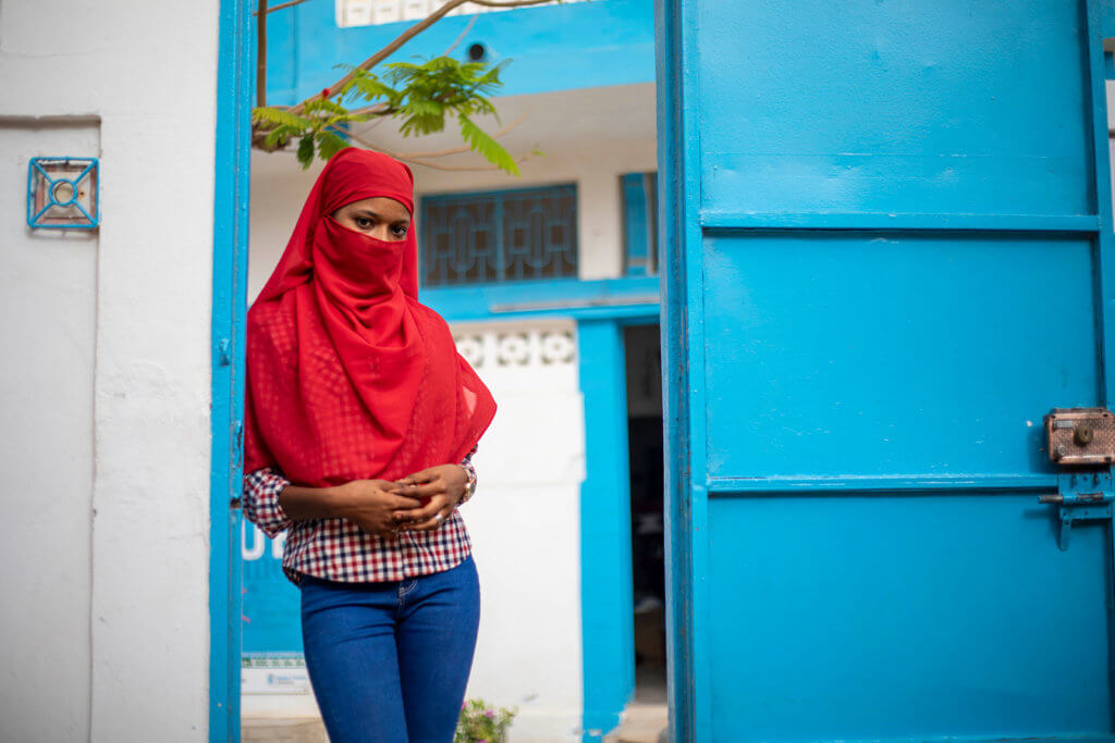 A young female student in a headscarf stands in front of an MSI center, with a blue-painted door.