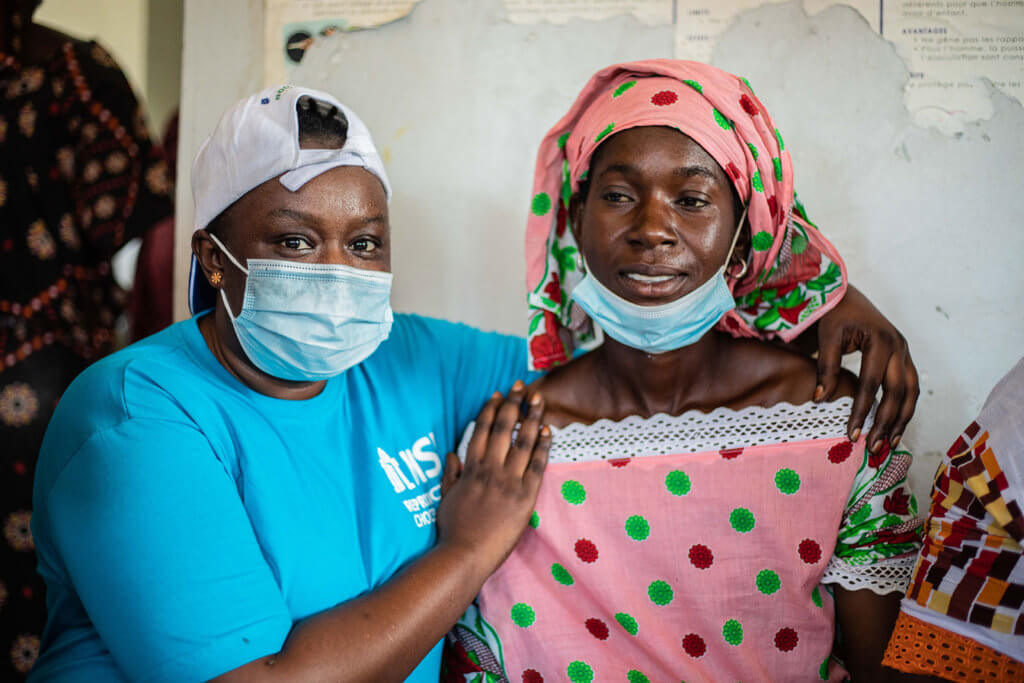 Fambaye, a woman in an MSI t-shirt wearing a surgical masks, sits with a client dressed in a pink head scarf and matching dress.
