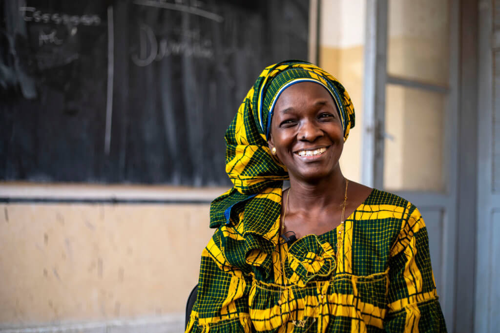 A Senegalese woman in a green and yellow dress sits in front of a blackboard, smiling.