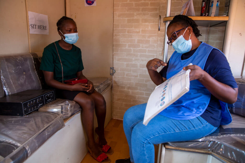 A young Zambian woman and an older woman sit in a small room. Both wear face masks. The older woman, a medical provider, is demonstrating different types of contraceptive.