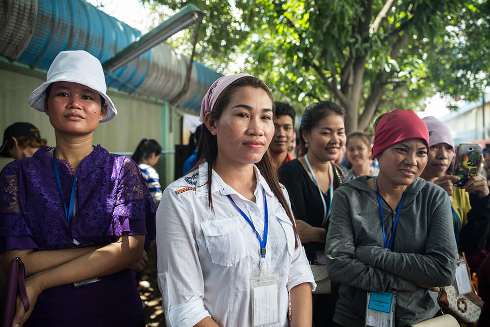 A group of Cambodian women, many wearing bandanas, smile outside a factory.