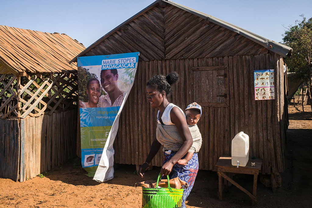 A young African woman wearing a baby on her back walks past a wooden building with an MSI poster outside.