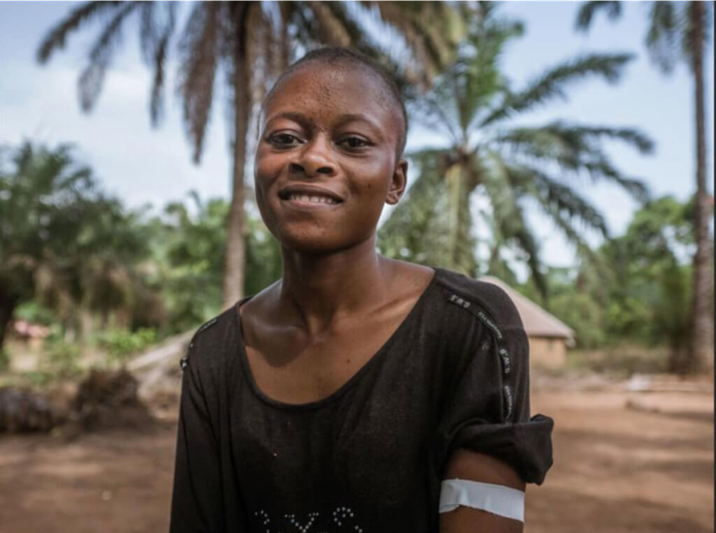 A young African woman looks at the camera. The background is a rural village. She has a bandage on her arm.
