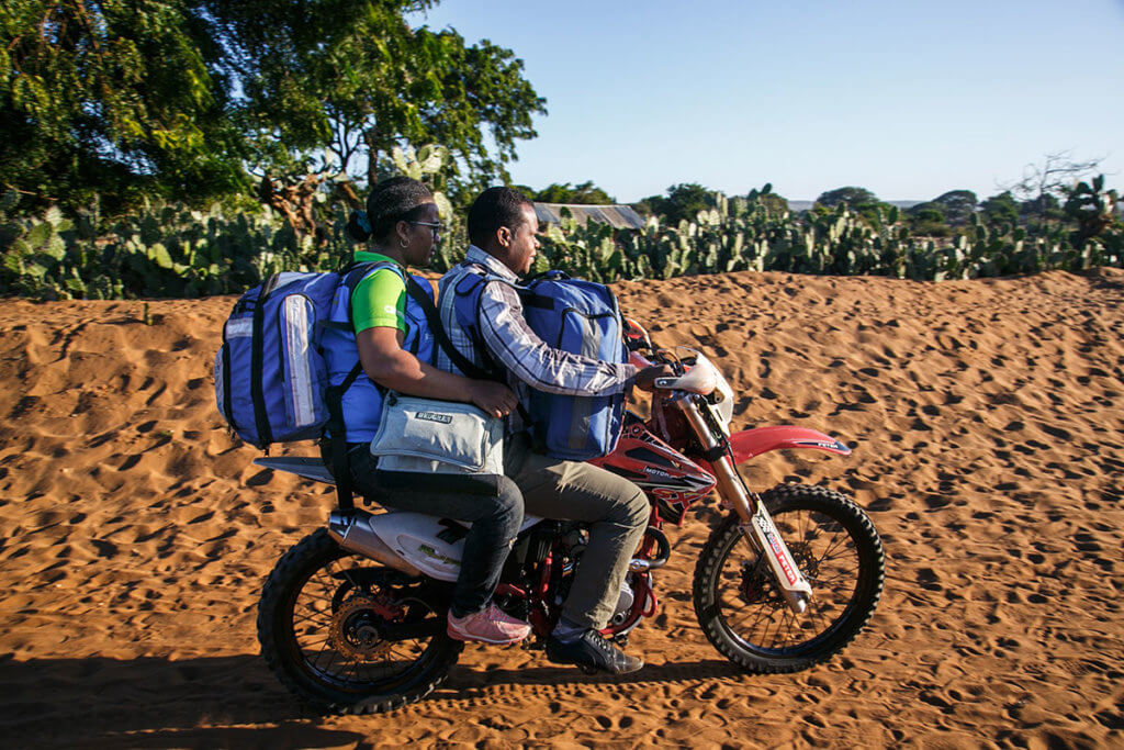 Jeannica, wearing a heavy backpack, rides a motorbike on a sandy road with cactuses in the background.