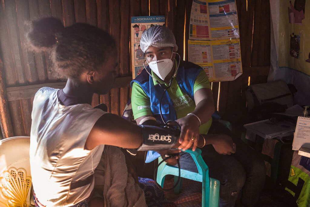 Jeannica, wearing a hair net and face mask, takes a woman's blood pressure in a dimly lit health clinic.