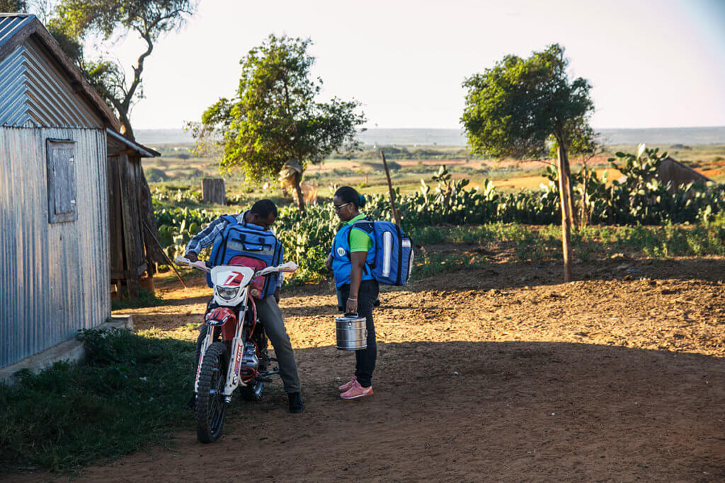 A man sits on a motorbike, wearing a large backpack. A woman stands next to him, also wearing a backpack and carrying medical supplies. They're traveling in a rural area of Madagascar on a dusty road.