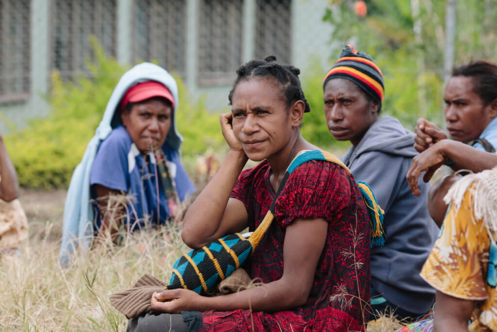 Women sit on the ground listening to a presentation at a rural health site.